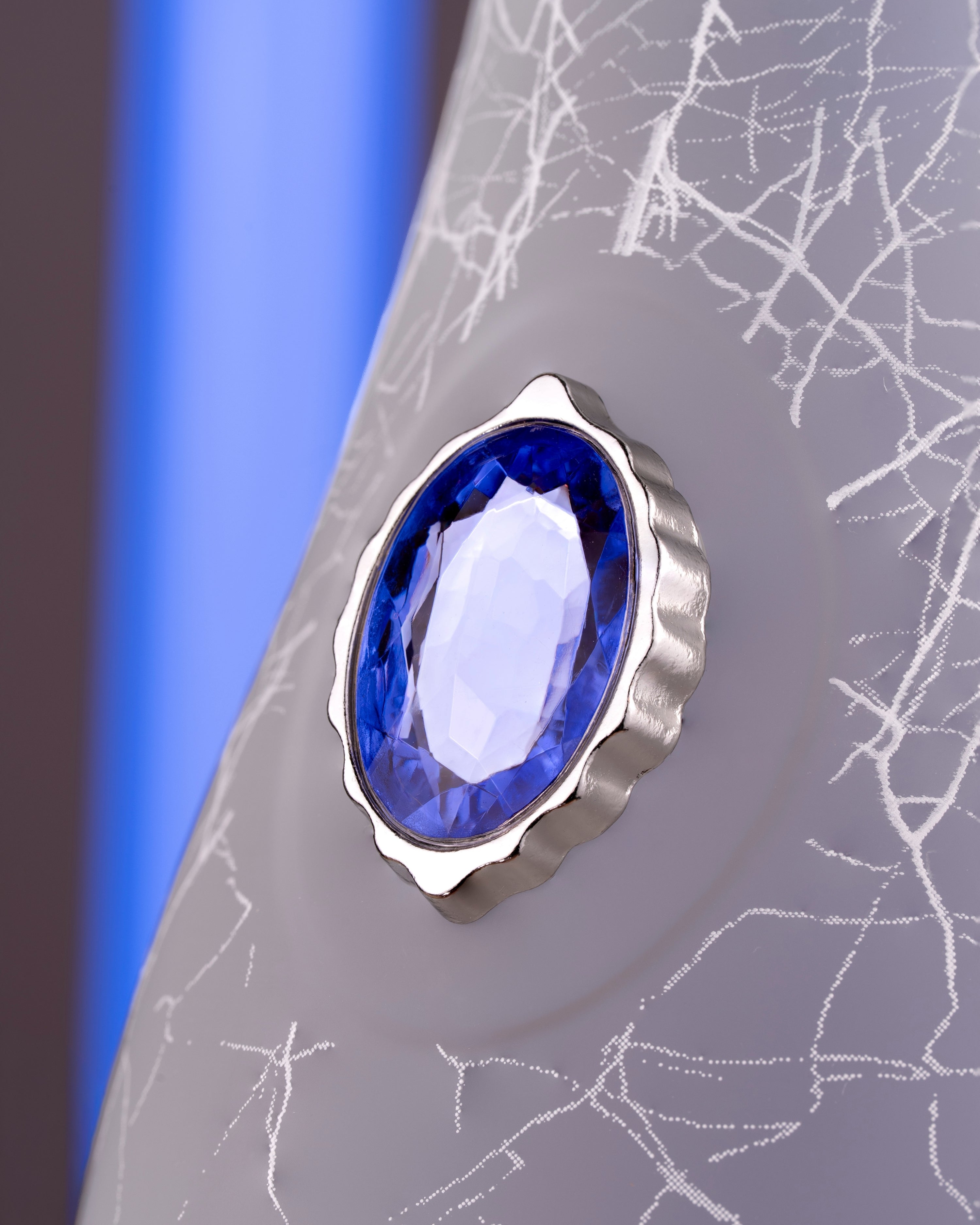 A close-up of a faceted oval blue gemstone set in a scalloped silver bezel, mounted on a textured gray decorative surface with white crackle-style lines.