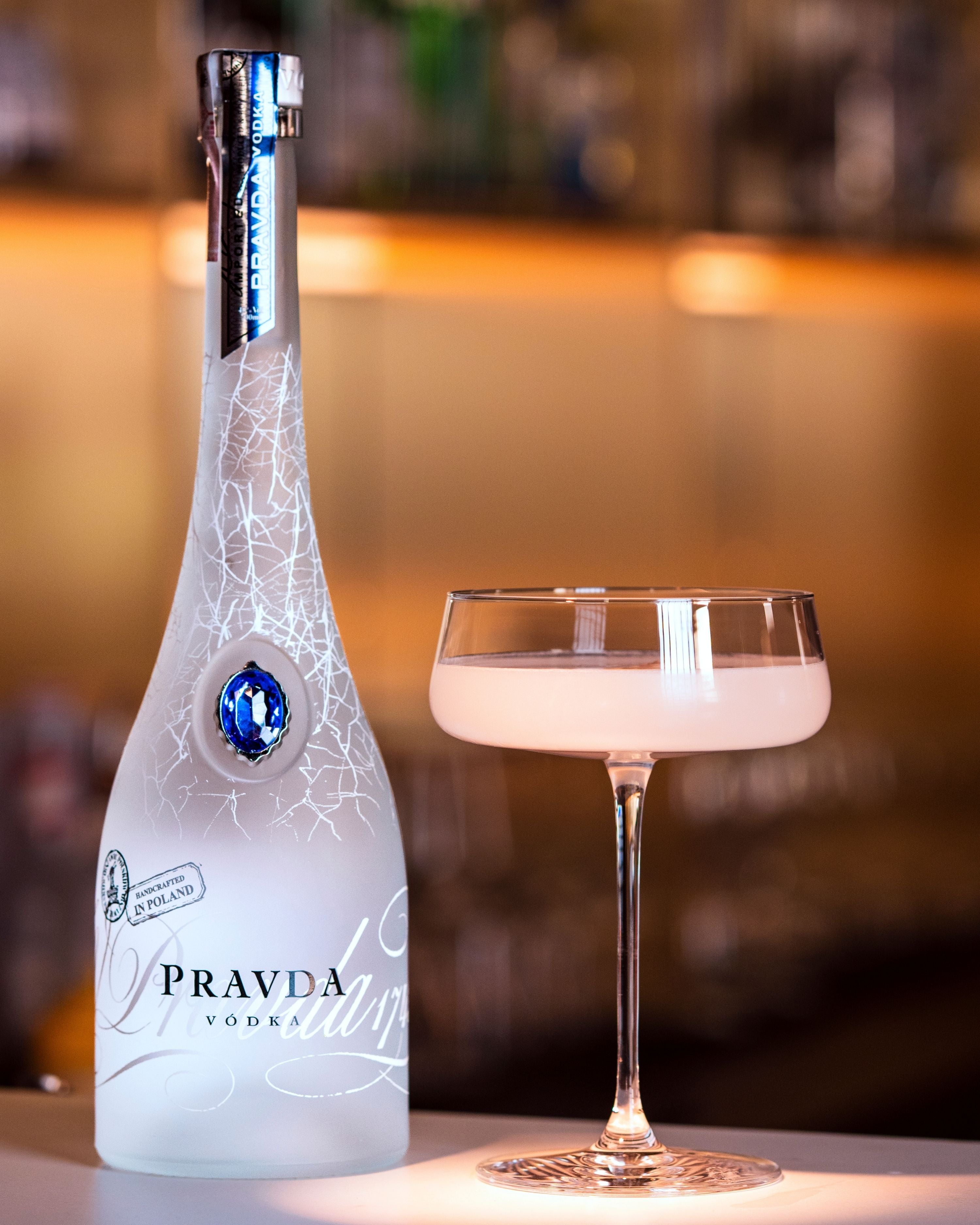 A bottle of Pravda Vodka next to a coupe glass filled with a pale drink, set against a softly blurred bar background.