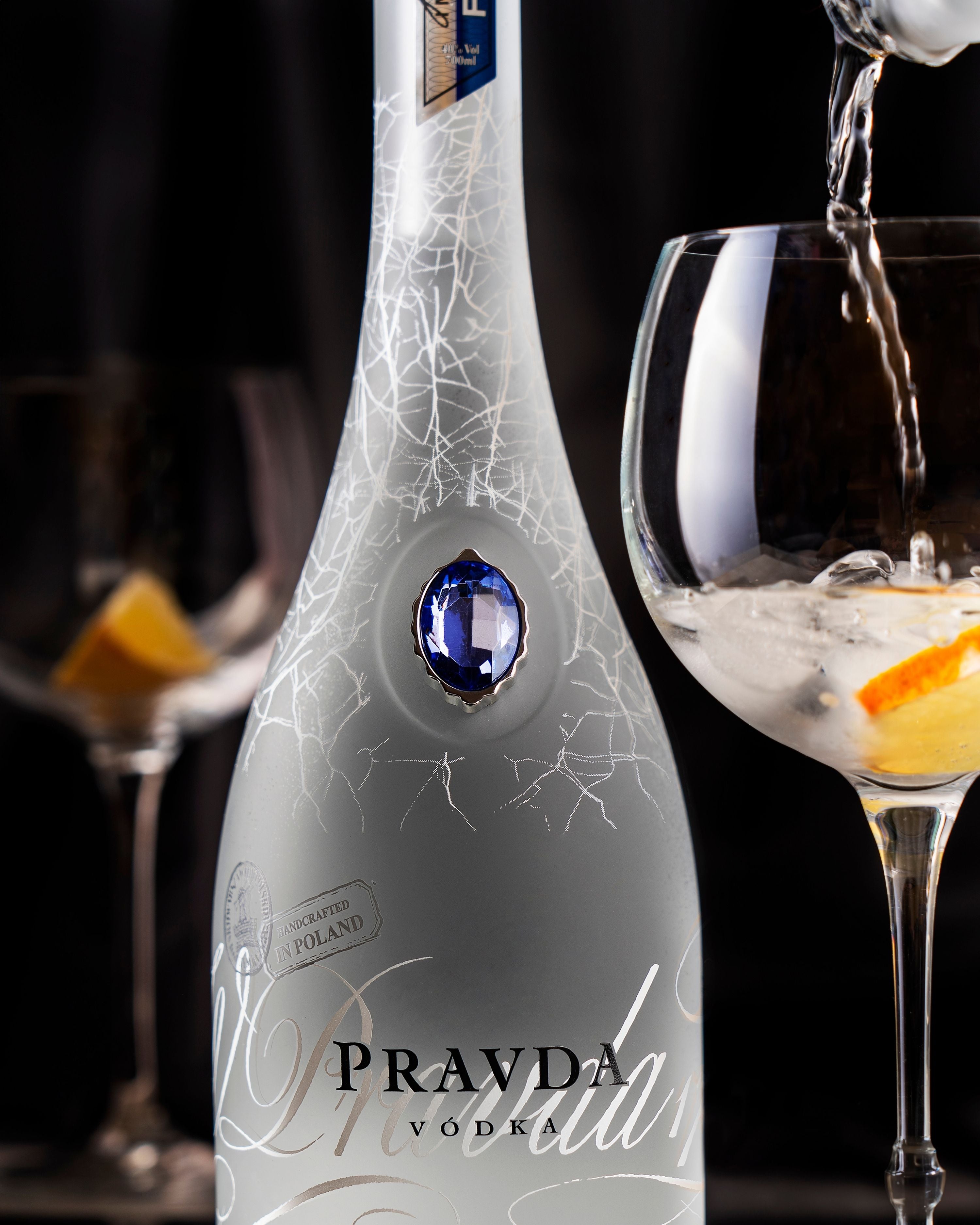 A bottle of Pravda Vodka with a silver-gray body, white crackle pattern, and a blue gem emblem, shown alongside two wine glasses, one of which has a clear liquid being poured into it.