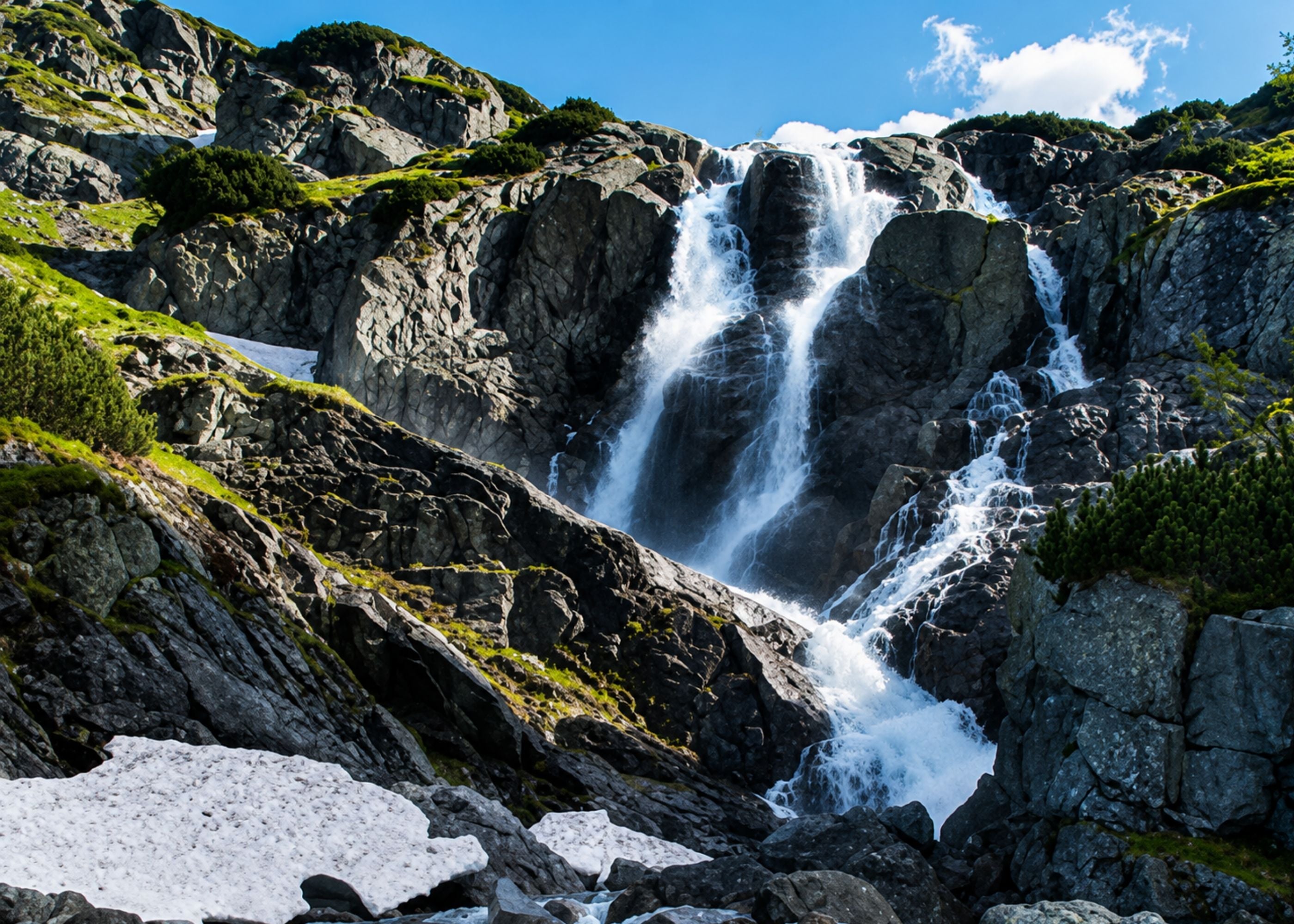 A scenic mountain landscape featuring a tall waterfall cascading down steep rocky cliffs, with patches of snow on the foreground and green vegetation on the rocks under a partly visible sky.