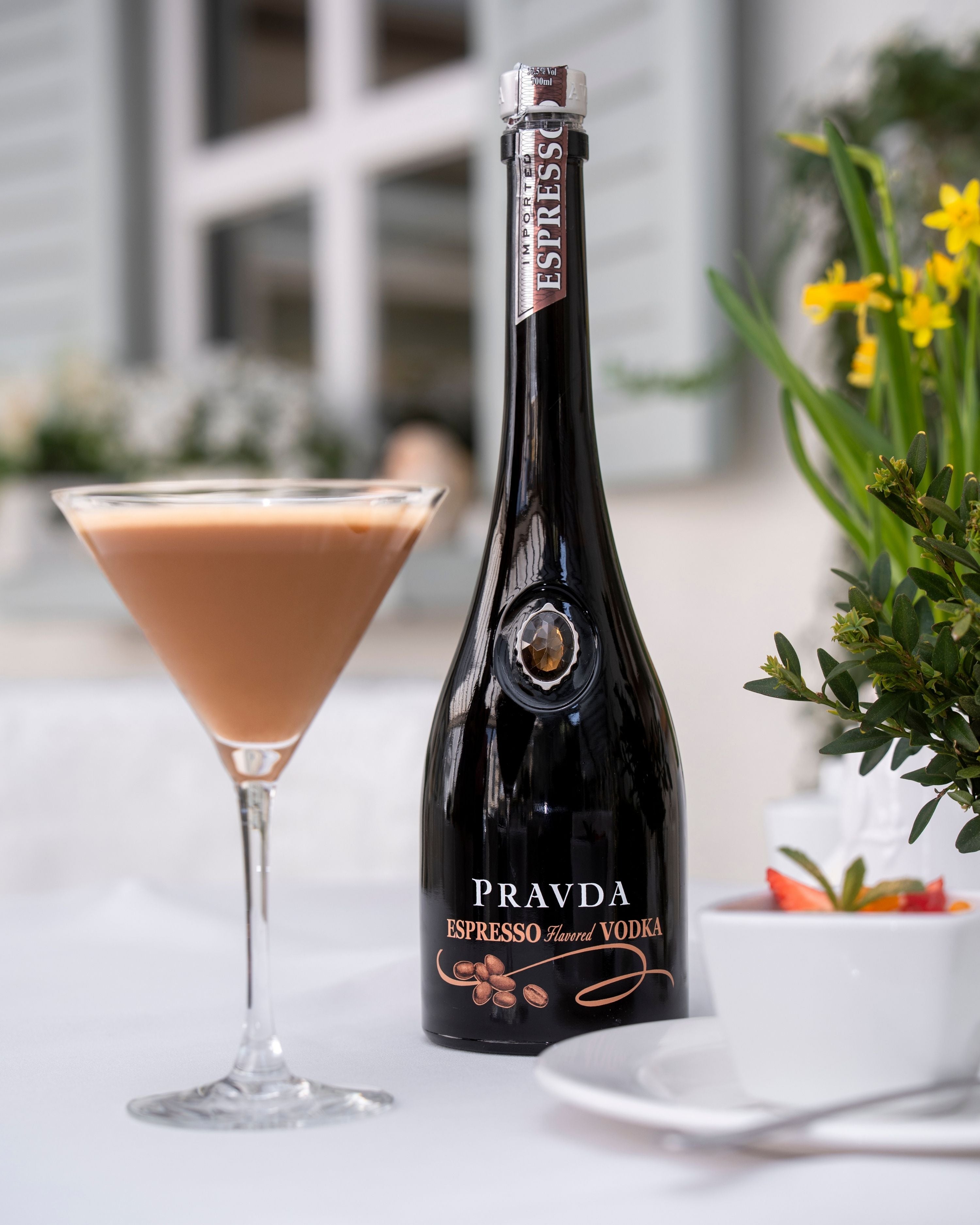 A bottle of Pravda Espresso Flavored Vodka stands on a white tablecloth beside a filled martini glass, with flowers and a cup in a softly blurred outdoor setting.