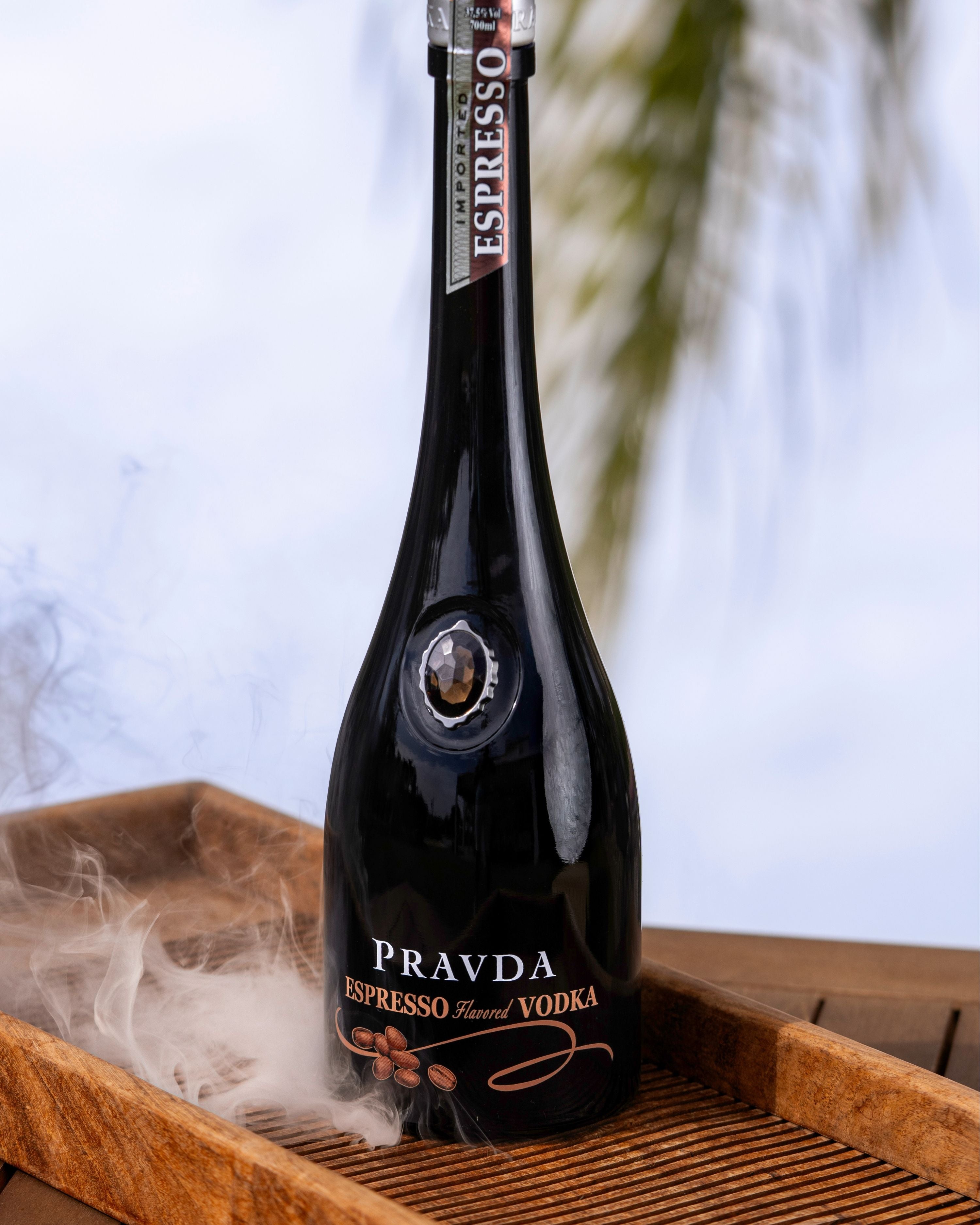 A bottle of Pravda Espresso Flavored Vodka styled on a wooden tray with a smoky effect, set against a soft-focus outdoor background with palm leaves.