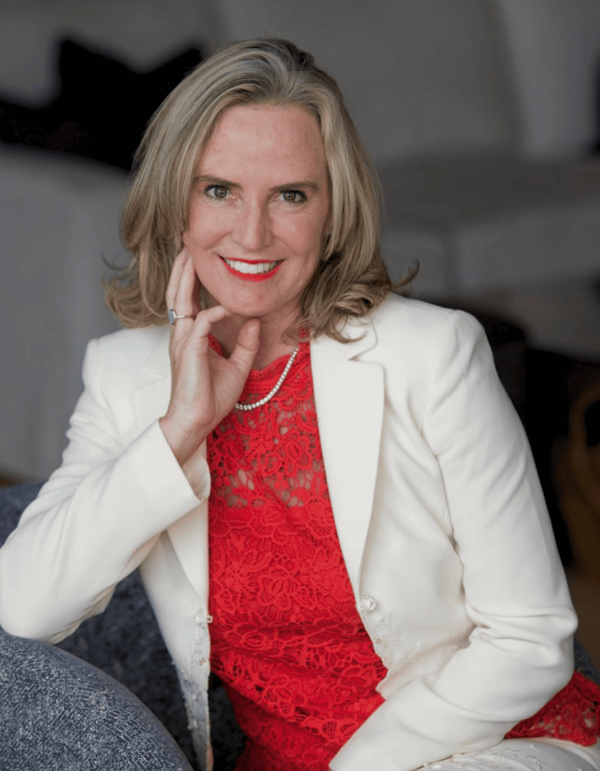 A smiling blonde woman posing indoors, wearing a white blazer over a bright red lace top, with a pearl necklace and a ring.