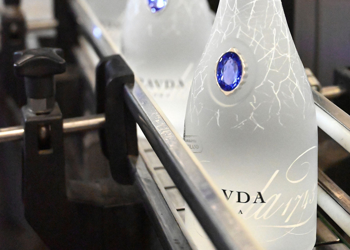 A frosted glass bottle with a blue oval gemstone and white crackle detailing, moving on a conveyor belt. The brand name 'PRAVDA' is partially visible on the bottle.