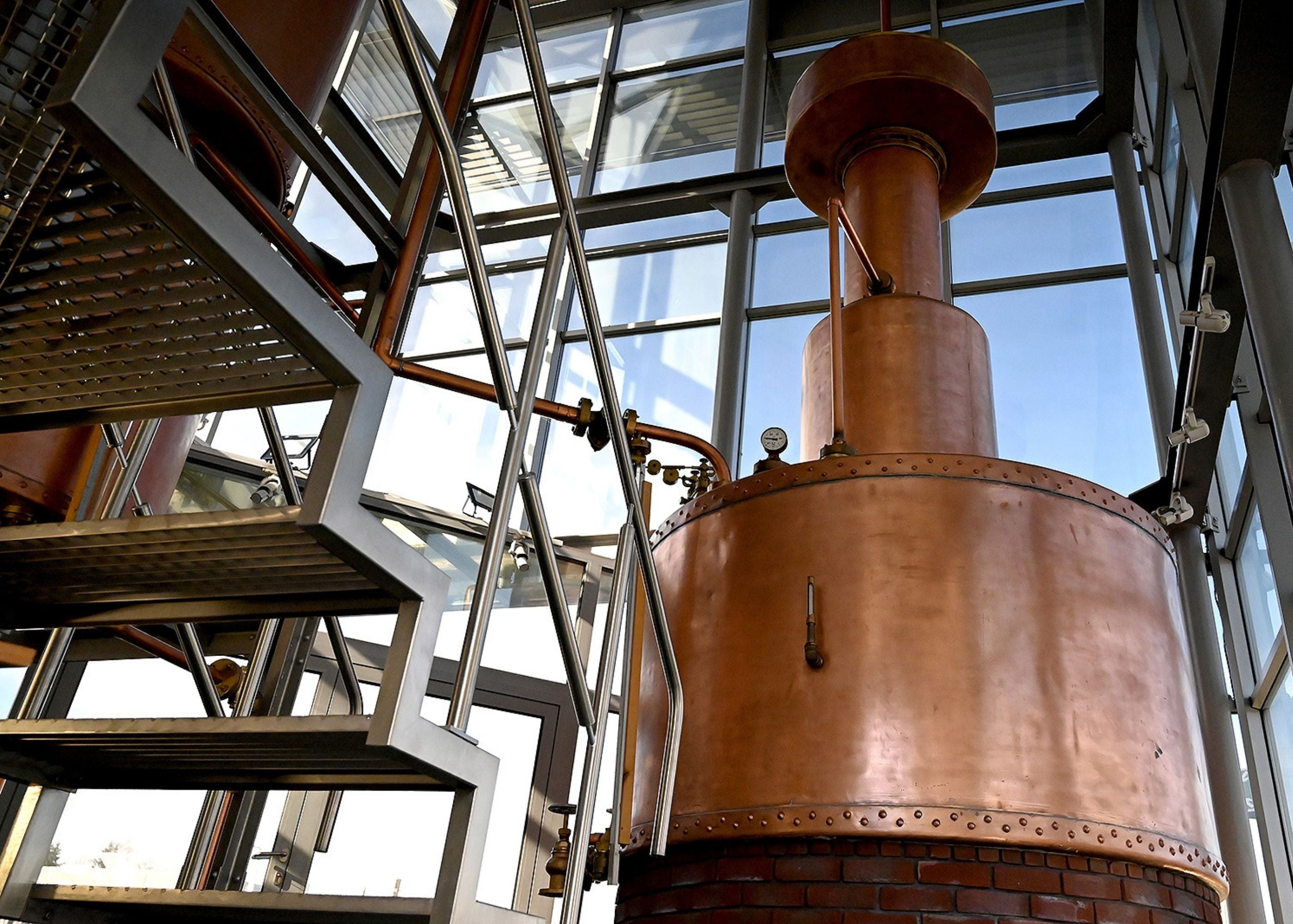 A large copper distillation still with metal stairs and glass windows in an industrial interior.