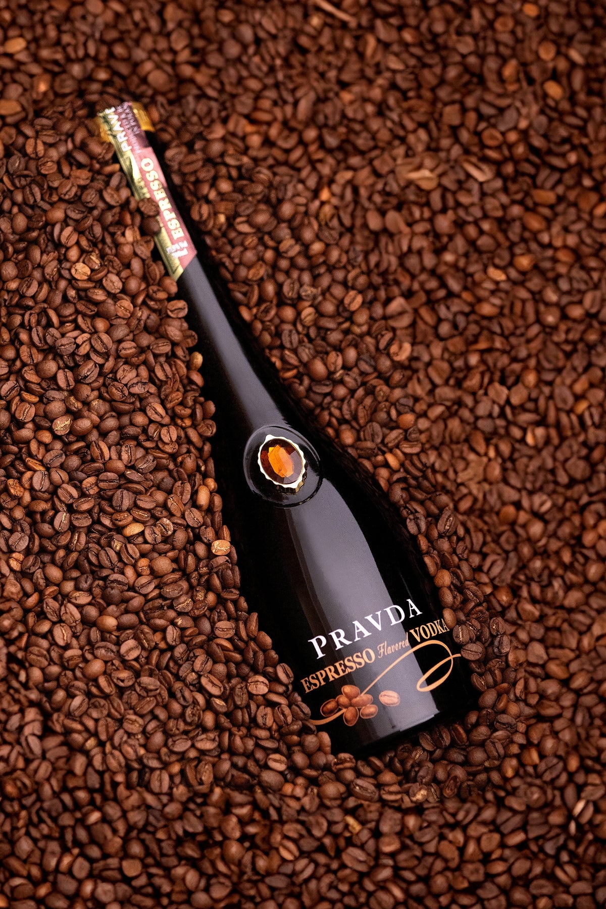 A bottle of PRAVDA Espresso Vodka lies diagonally on a dense bed of roasted coffee beans. The bottle is dark glass with gold lettering and a gold seal near the neck.