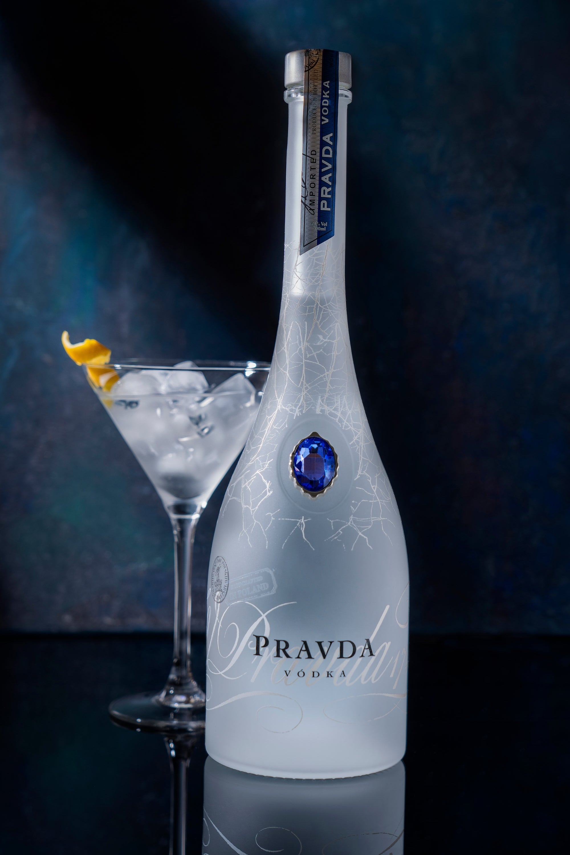 A bottle of Pravda Vodka with a blue gemstone accent and frosted glass, shown next to a martini glass filled with ice and a lemon twist, on a dark reflective surface against a dark blue background.