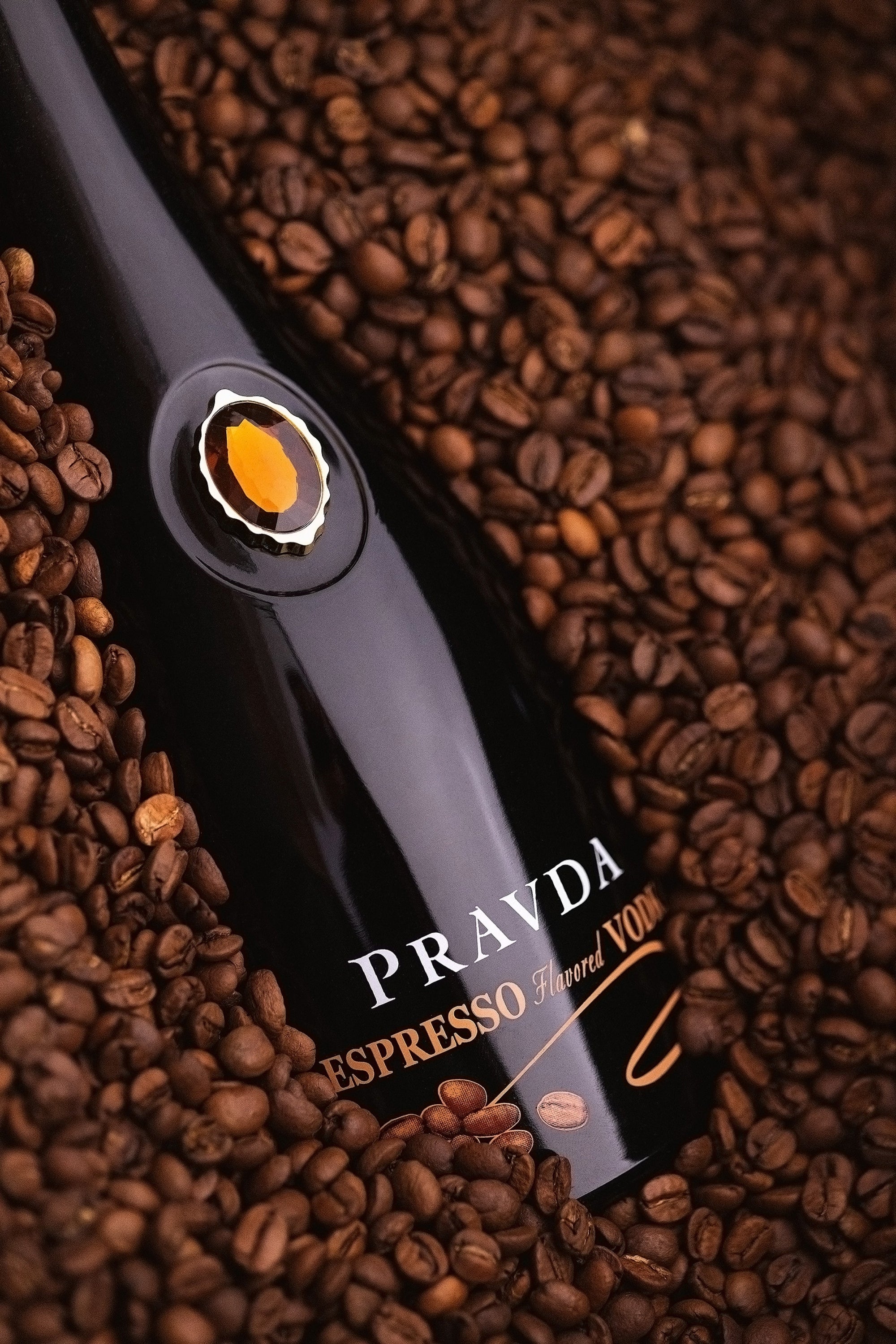 A bottle of Pravda Espresso flavored Vodka nestled among scattered roasted coffee beans. The bottle is black with gold and white lettering, and a gold and black label.