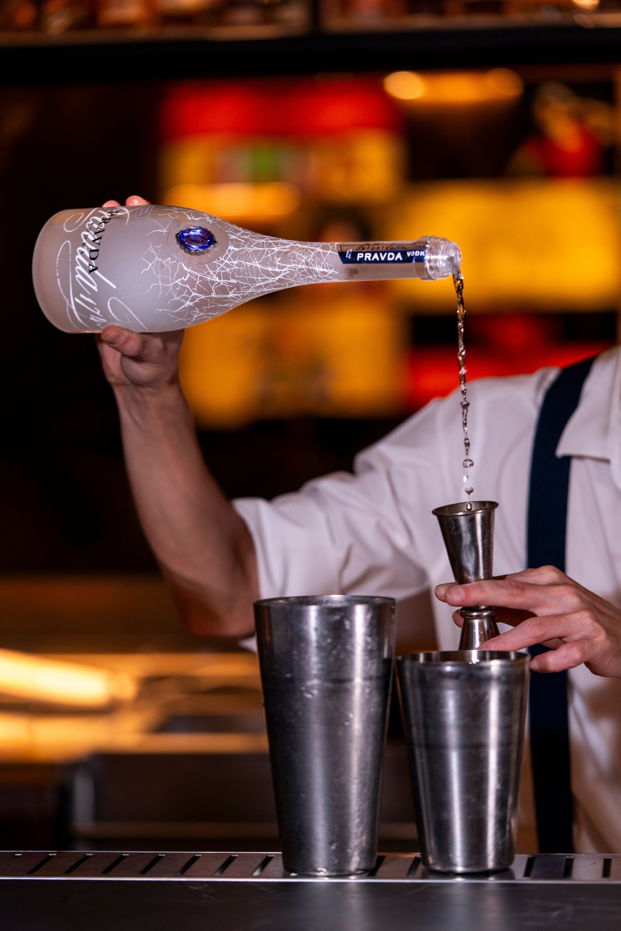 A bartender in a white shirt and dark suspenders pours a clear liquid from a frosted glass bottle labeled 'PRAVDA' into metal jigger on a bar counter, with a warm, blurred bar background.