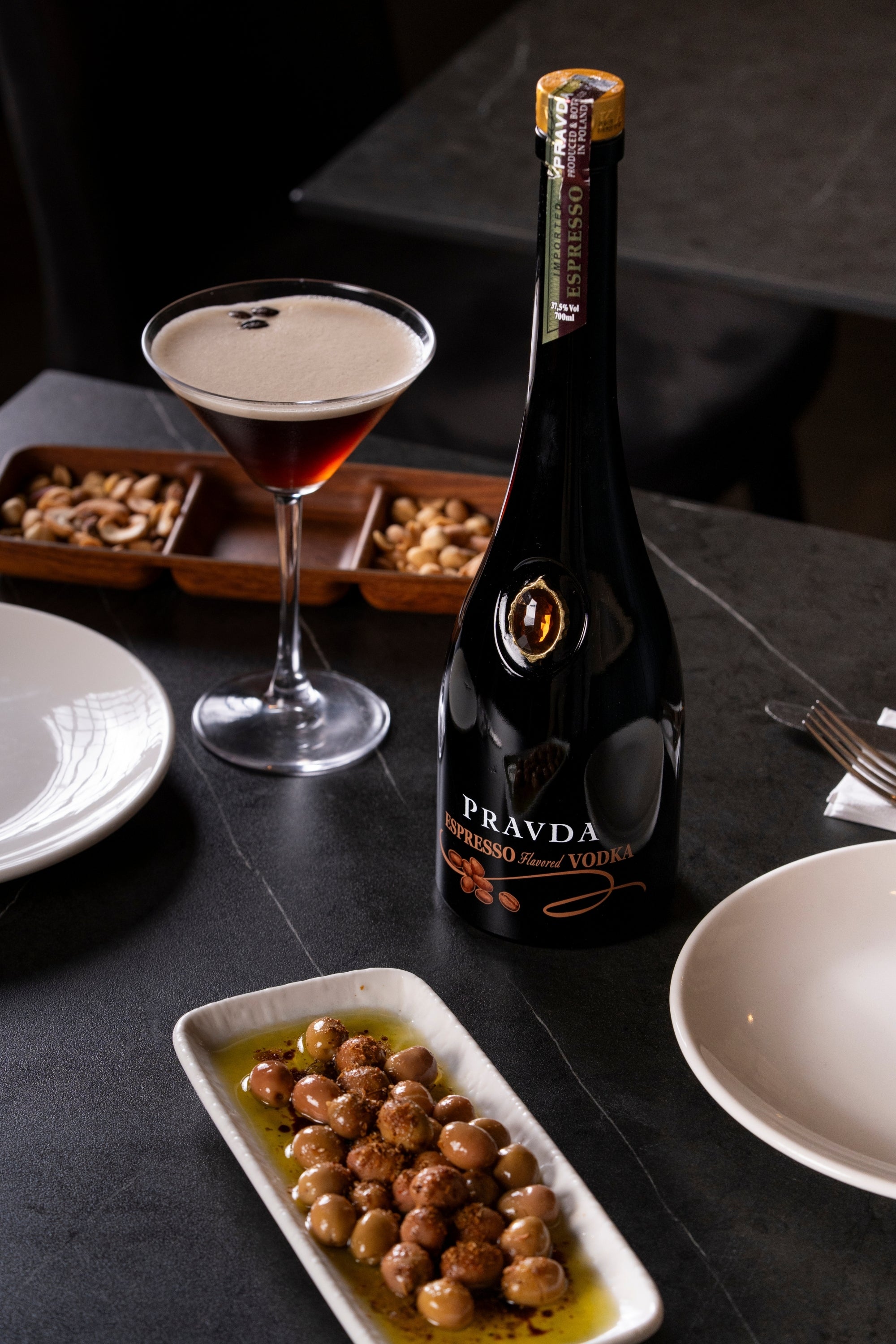 A bottle of Pravda Espresso Vodka stands on a dark tabletop beside a filled martini glass, plates, and small dishes of nuts and olives in a dimly lit restaurant setting.