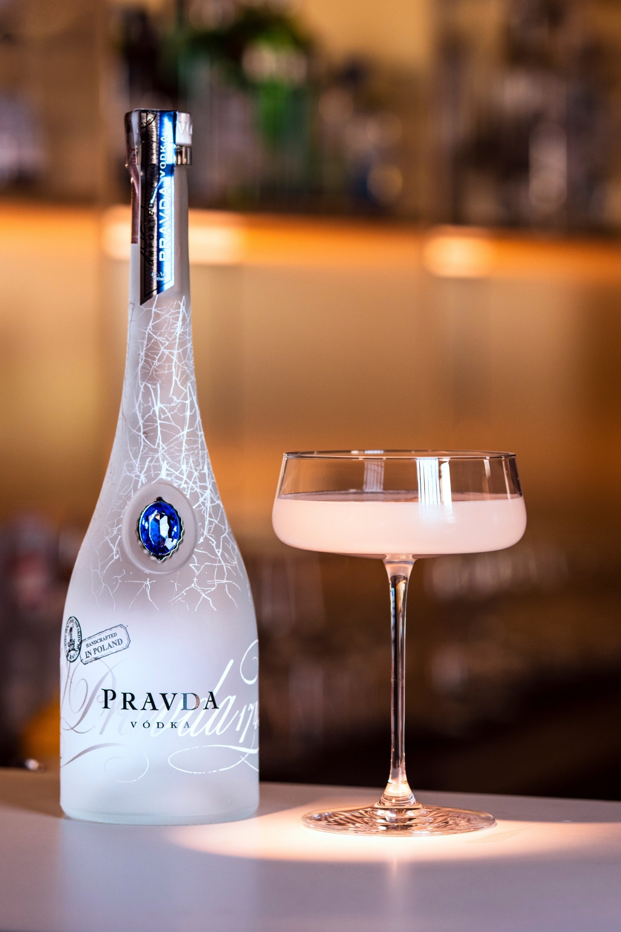 A bottle of Pravda Vodka next to a coupe glass filled with a pale pink cocktail, set on a bar counter with a blurred bar background.