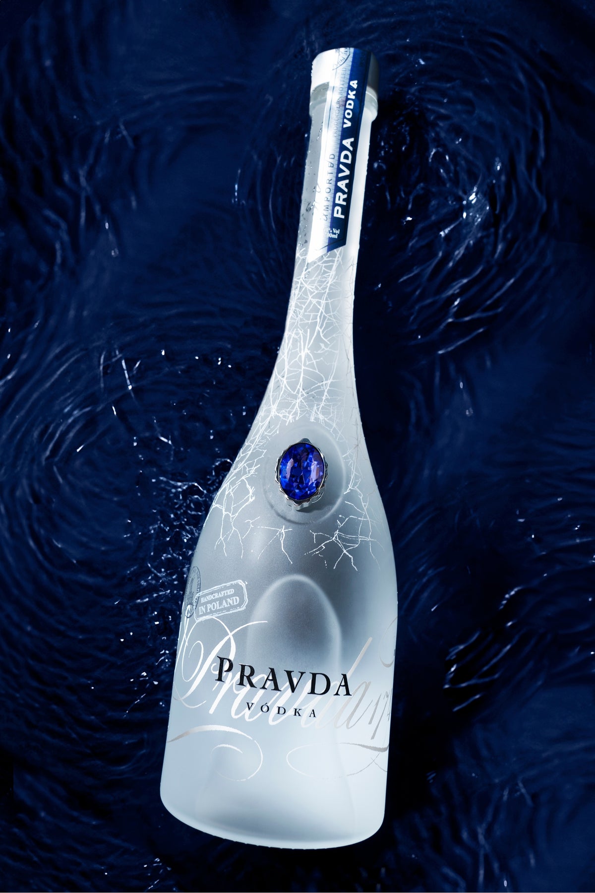 A frosted glass bottle of Pravda Vodka with a blue gemstone and white crackle-style etched pattern, set against a dark blue water-textured background.