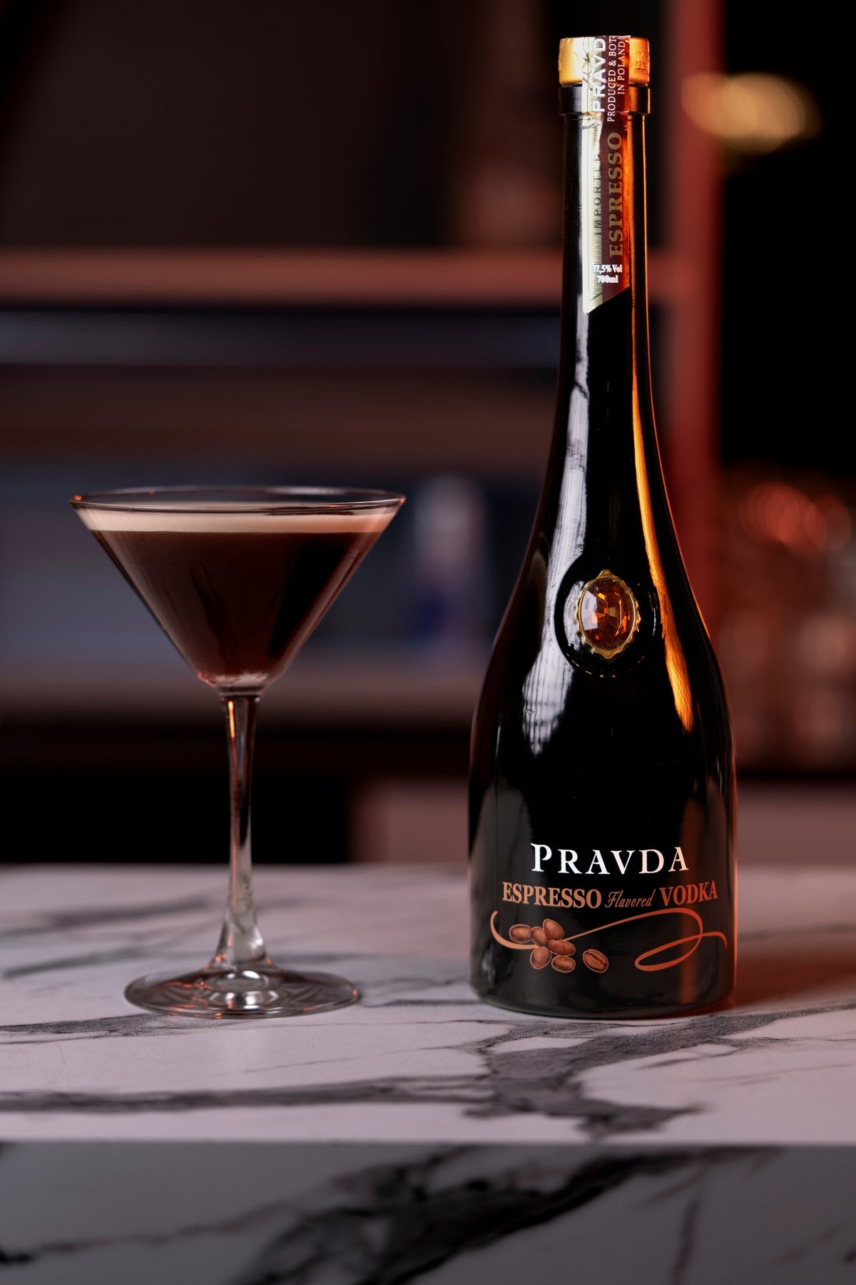 A bottle of Pravda Espresso Vodka stands on a marble surface next to a filled martini glass with a dark reddish-brown drink, set against a softly blurred bar background.