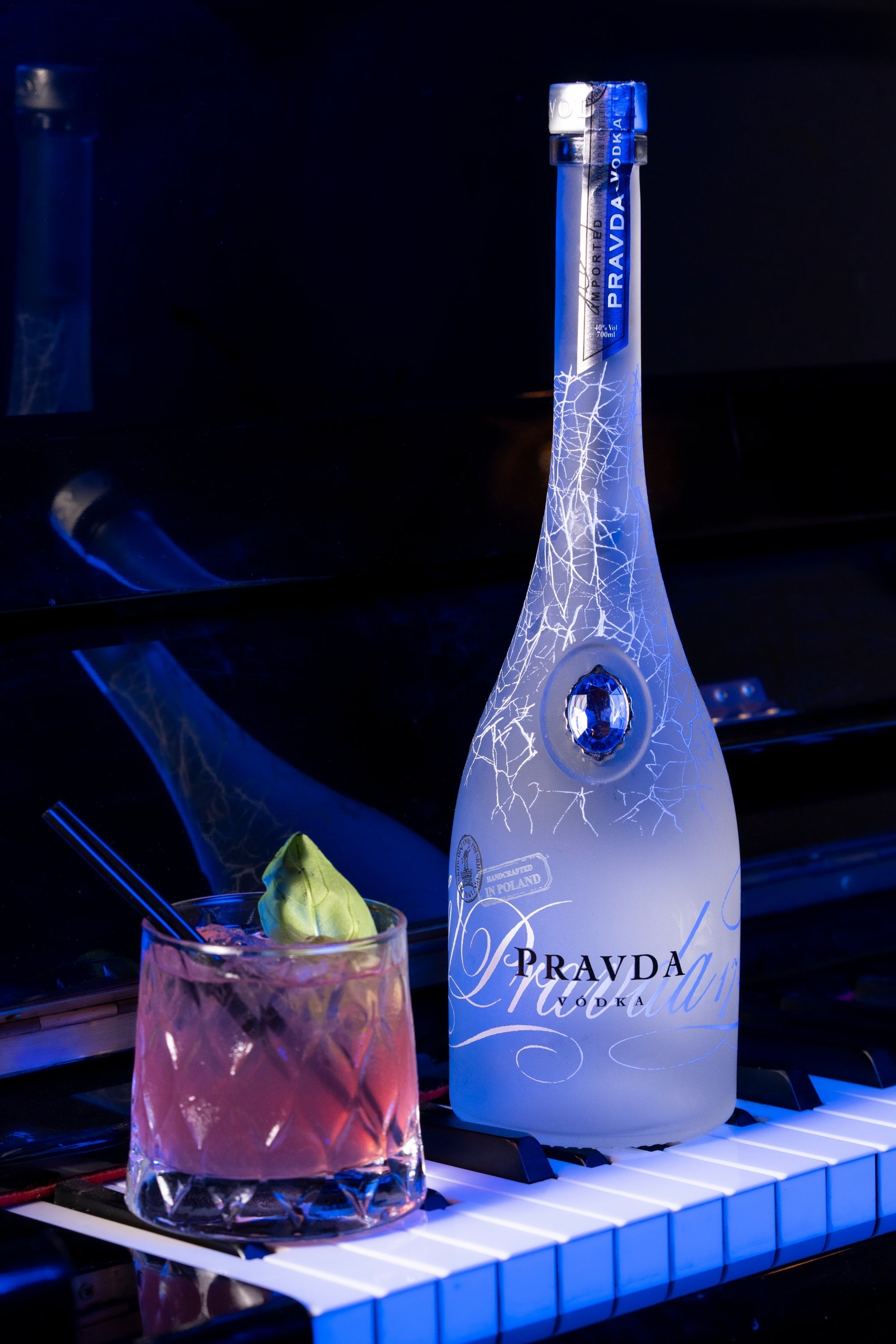 A bottle of Pravda Vodka with white decorative script and crackle patterns, shown beside a pink cocktail in a glass on a piano keyboard against a dark blue background.