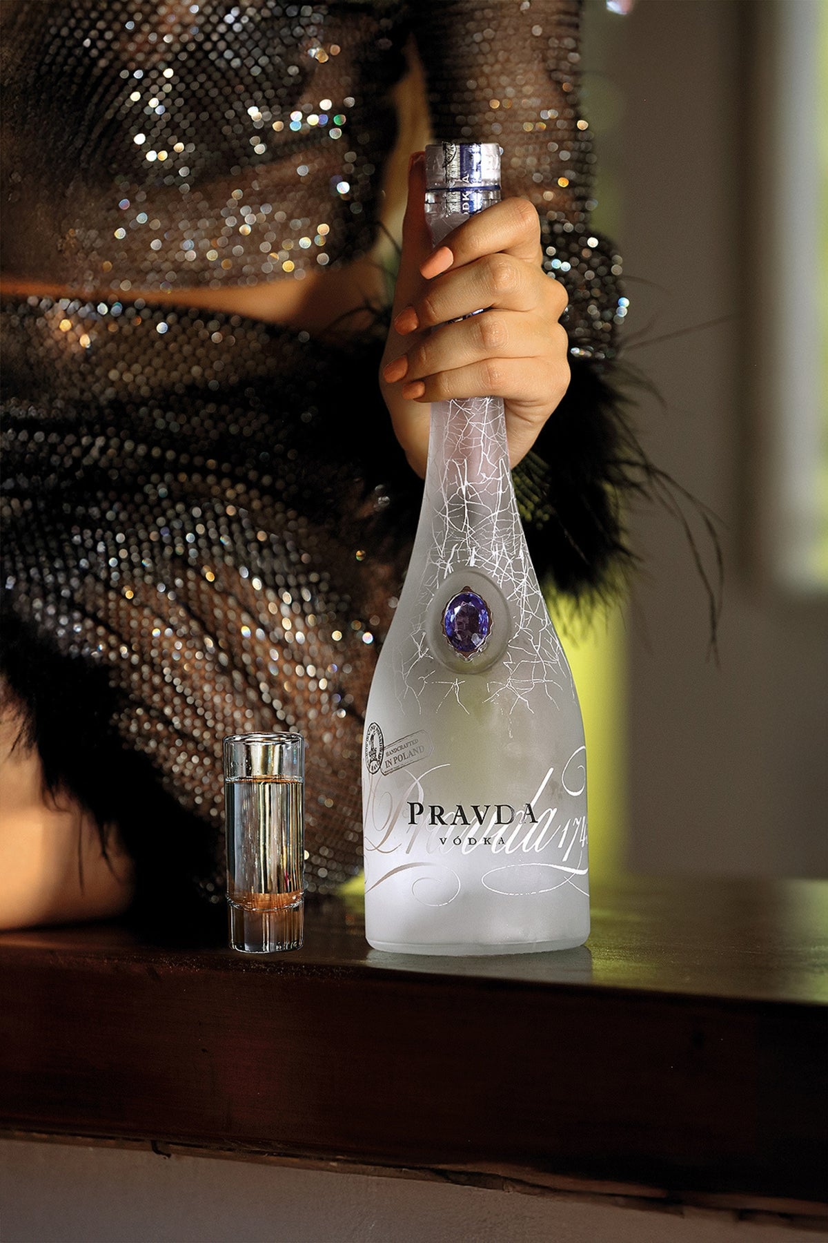 A frosted glass bottle of Prada Vodka with a blue crystal seal and silver script, held by a person wearing a sparkling black dress, with a small silver tumbler on a dark wooden surface.