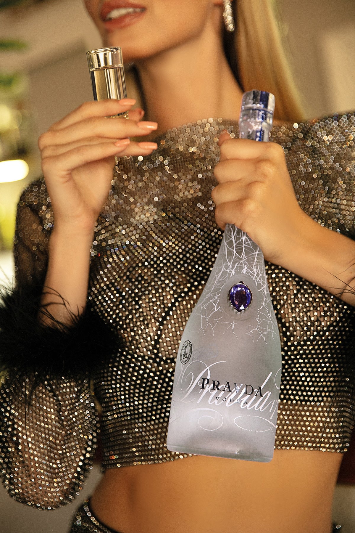 A woman in a sparkling silver sequined crop top holds a frosted glass bottle of Pravda vodka with a blue gemstone and silver foil, while another hand holds a small shot glass.
