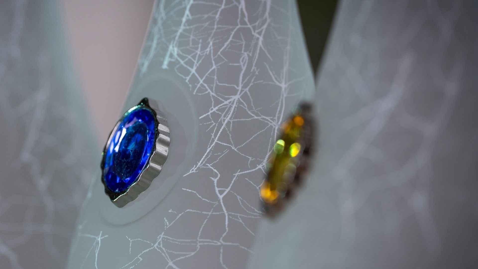 A close-up of a sculptural, textured white bottle featuring a prominent oval blue crystal signet on the left and a blurred amber crystal signet on the right.