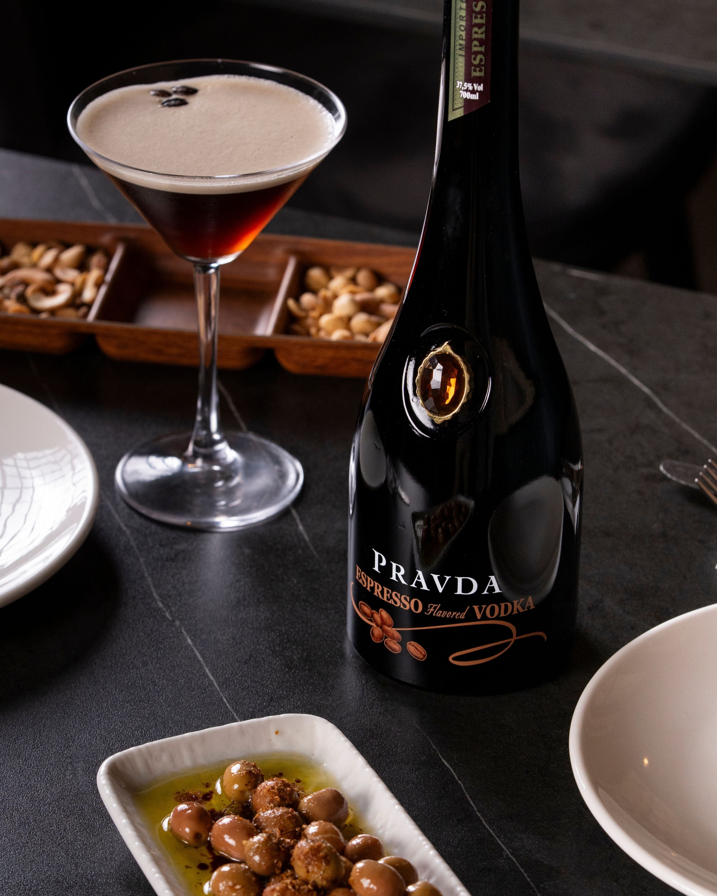 A bottle of Pravda Espresso Flavour Vodka stands on a dark tabletop beside a martini glass filled with a dark red drink and foamy head, with small dishes of nuts and olives in the background.