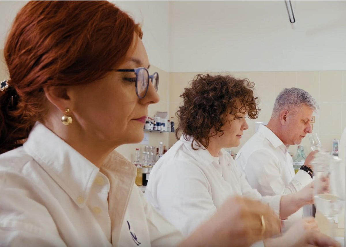 Three people in white lab coats working in a bright laboratory setting, with one woman in the foreground wearing glasses and red hair.