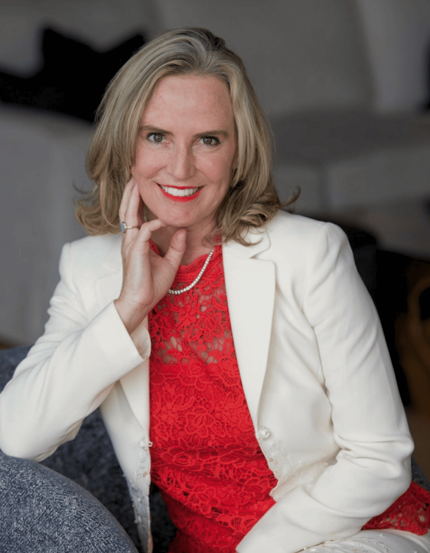 A smiling blonde woman posing indoors, wearing a white blazer over a bright red lace top, with a pearl necklace and a ring.