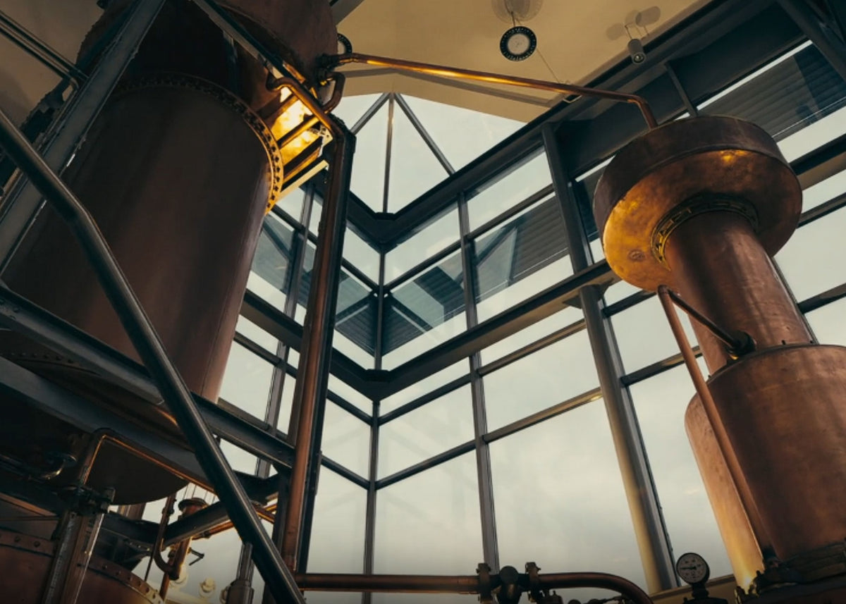 Industrial distillation apparatus with large copper columns and metal piping inside a glass-walled structure, photographed from a low angle.